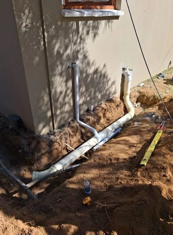 Exposed drainage pipework in a trench beside a building with vertical pipe connections